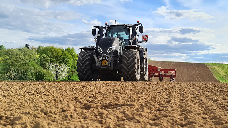 A large black Valtra T Series tractor is prominently featured in a freshly tilled field under a partly cloudy sky. The scene is set in a rural agricultural landscape with green trees and rolling hills in the background. The tractor's robust tyres and attached farming equipment are clearly visible, emphasising modern farming technology. 
