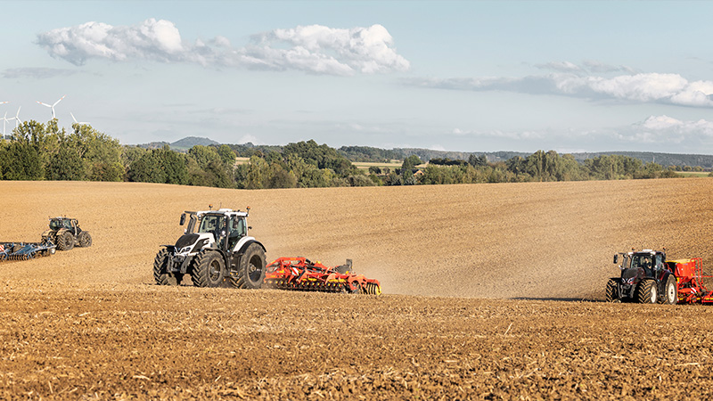 Trzy traktory z dołączonymi maszynami rolniczymi w trakcie prac na polu.